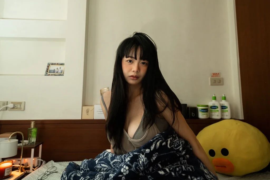 Person sitting on a bed in a lived-in bedroom, wearing casual home clothing.