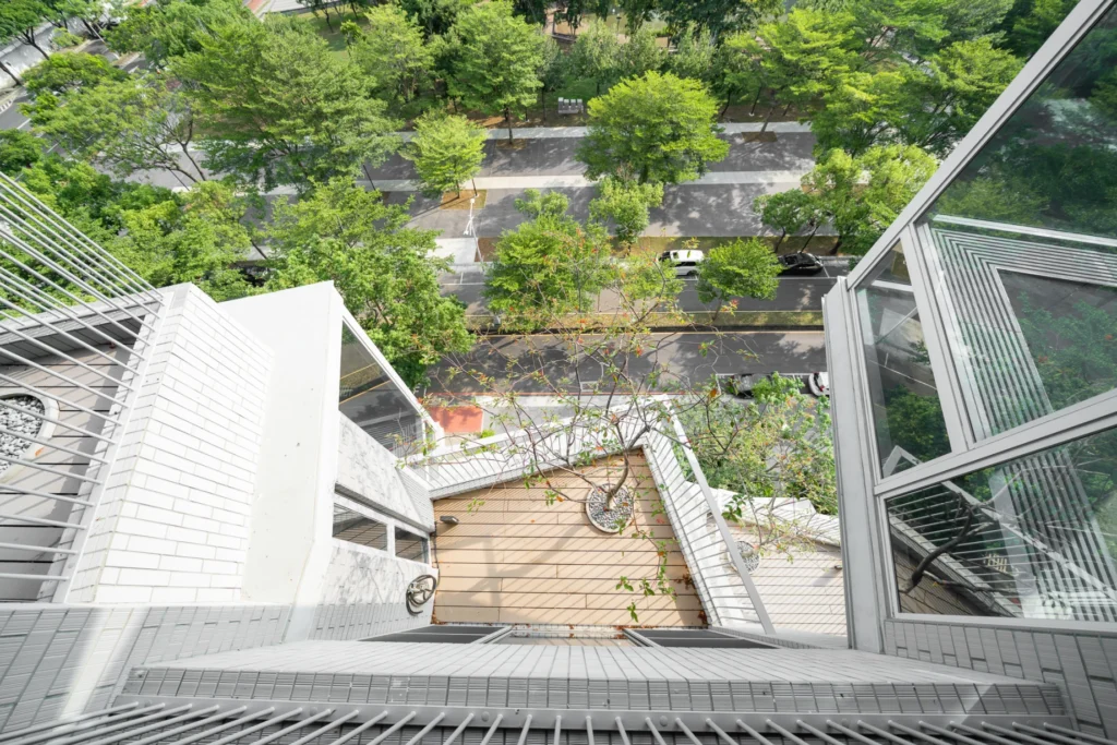 A downward view across staggered white terraces and railings, a tree on a timber-coloured deck, reflective glazing, and the tree-lined street below.