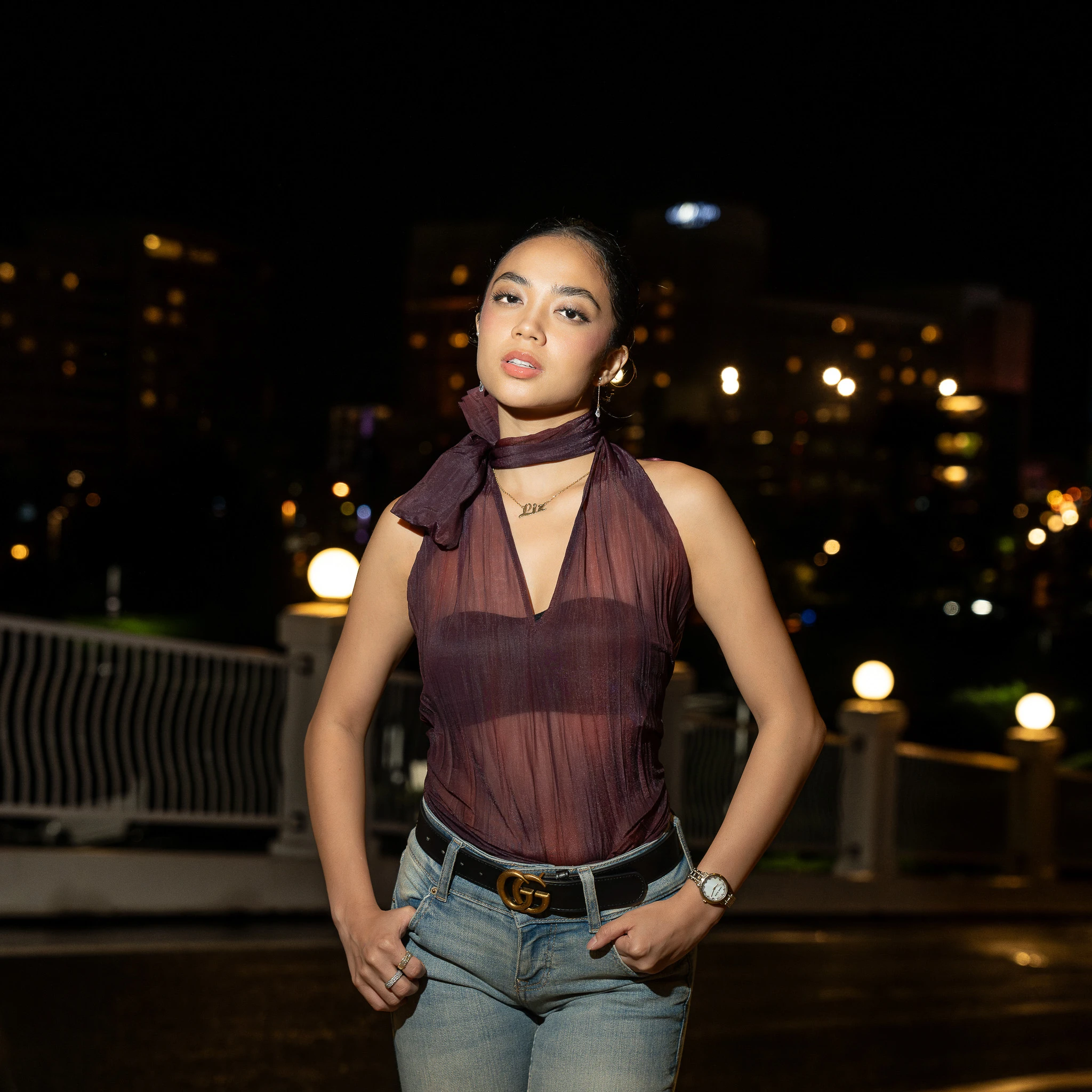 The subject stands by a railing at night, with round lamps and distant points of light behind her.