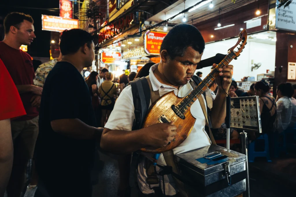 Street musician playing a stringed instrument among evening market visitors.