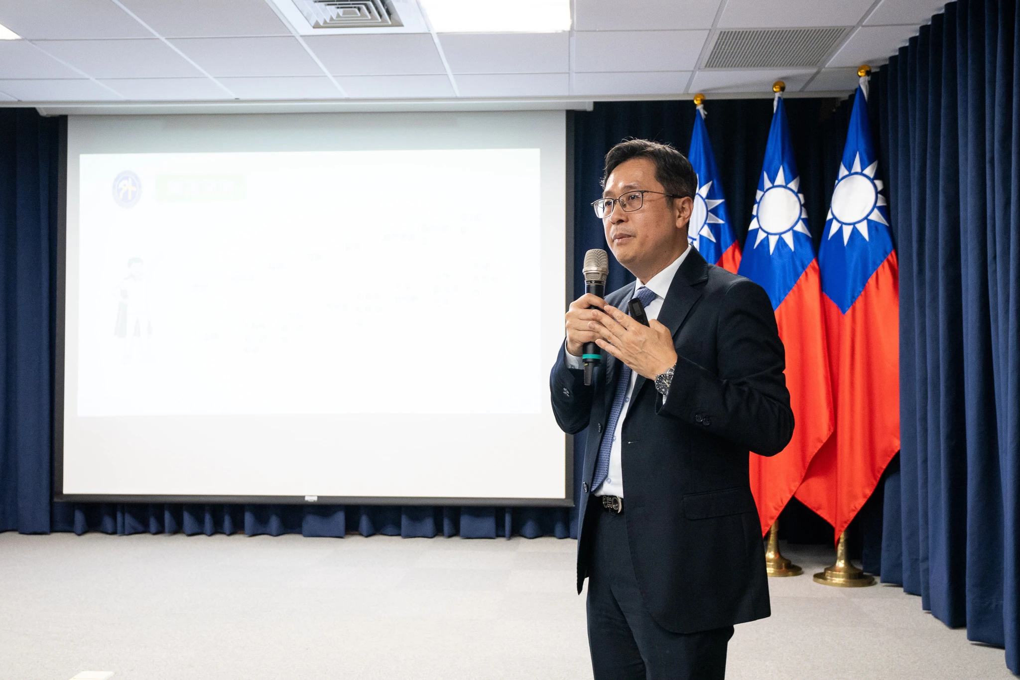 A suited speaker holds a microphone beside a projection screen and flags.