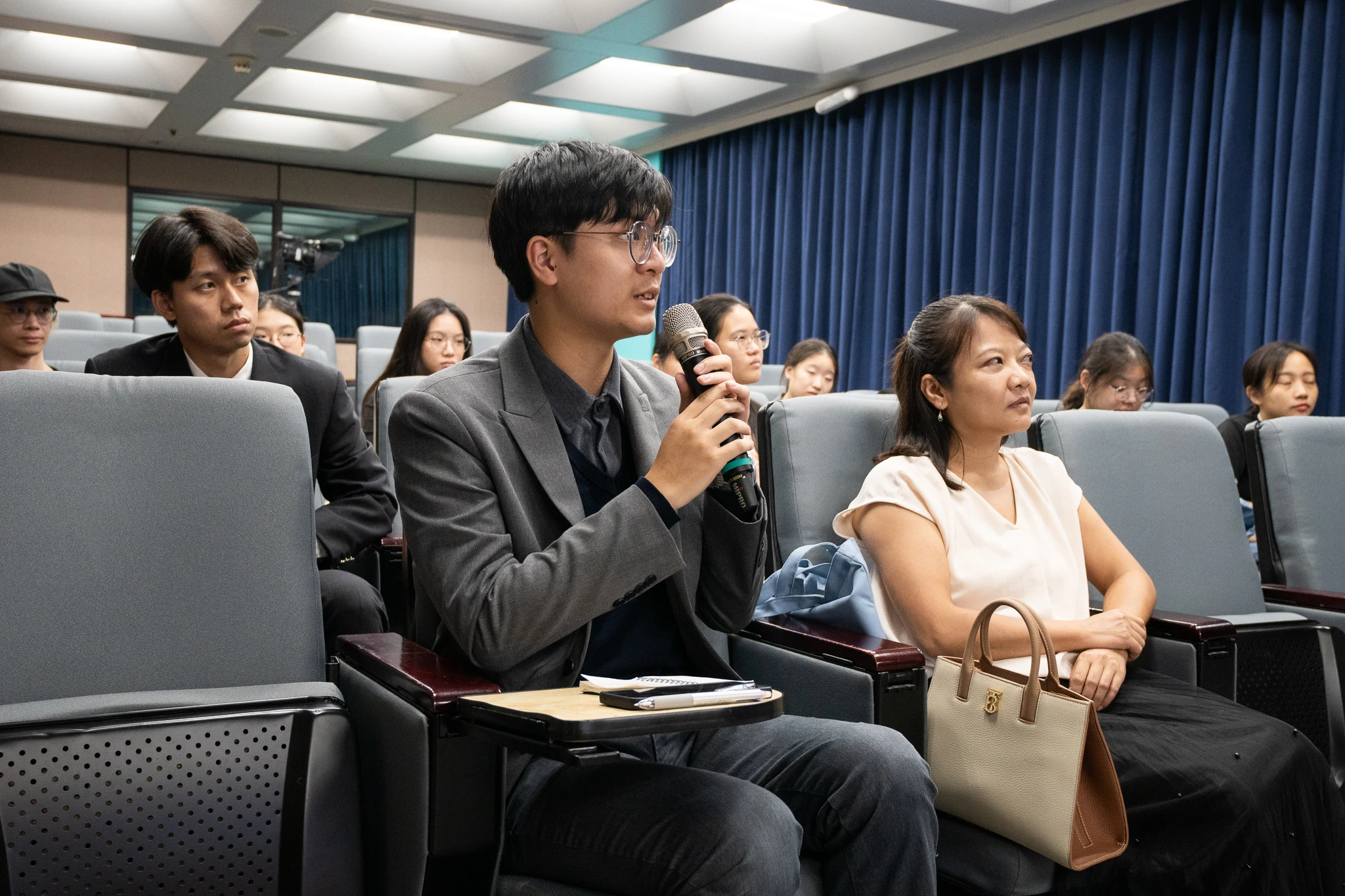 A seated participant speaks into a microphone, with other attendees and rows of chairs around him.