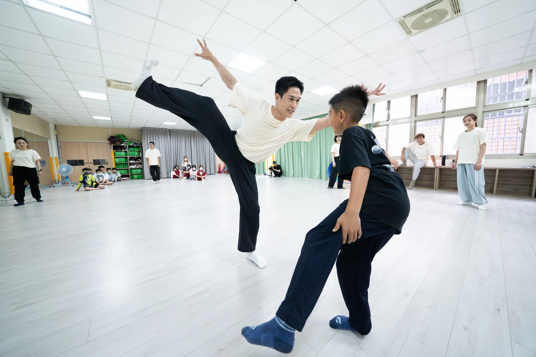 Two students practising a high kick and crouching movement in a rehearsal room.