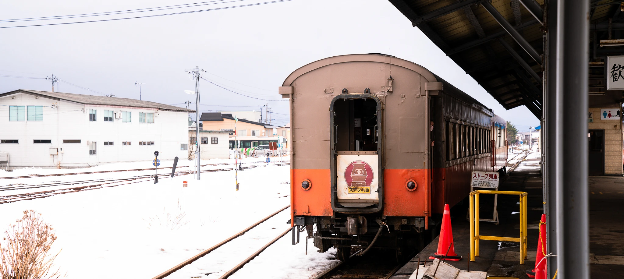 The brown and orange Tsugaru train stands beside a platform, with snow covering the tracks and nearby ground.
