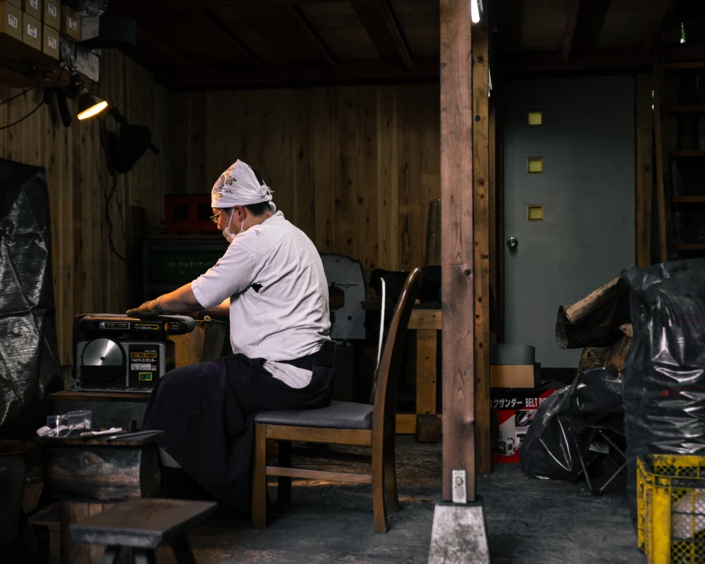 A craftsperson seated at a workbench inside a traditional metal workshop.