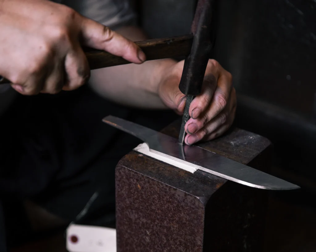 Hands hold a wooden-handled hammer and a narrow tool, its tip resting against a knife blade on a metal block.